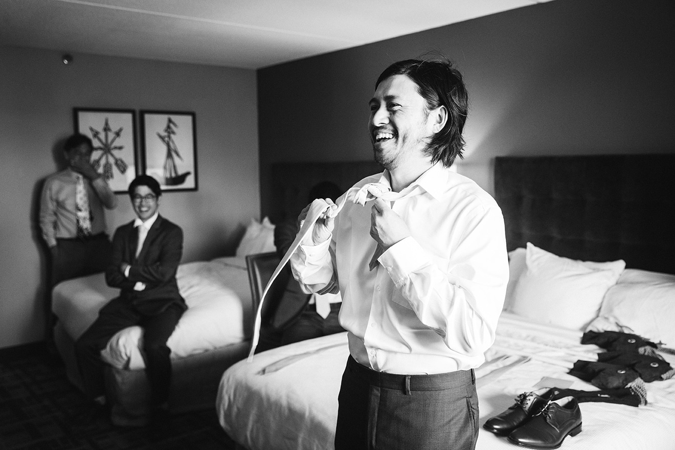 A documentary photograph of a groom laughing while he tying his tie before his plimoth plantation wedding