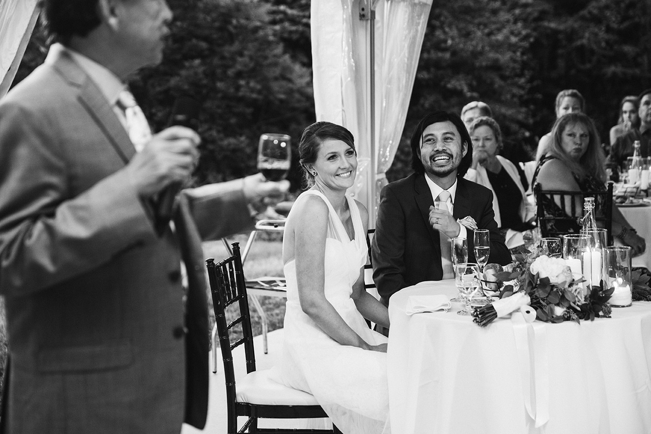 A documentary photograph of a bride and groom smiling during the father of the groom toast at their plimoth plantation wedding in plymouth, massachusetts