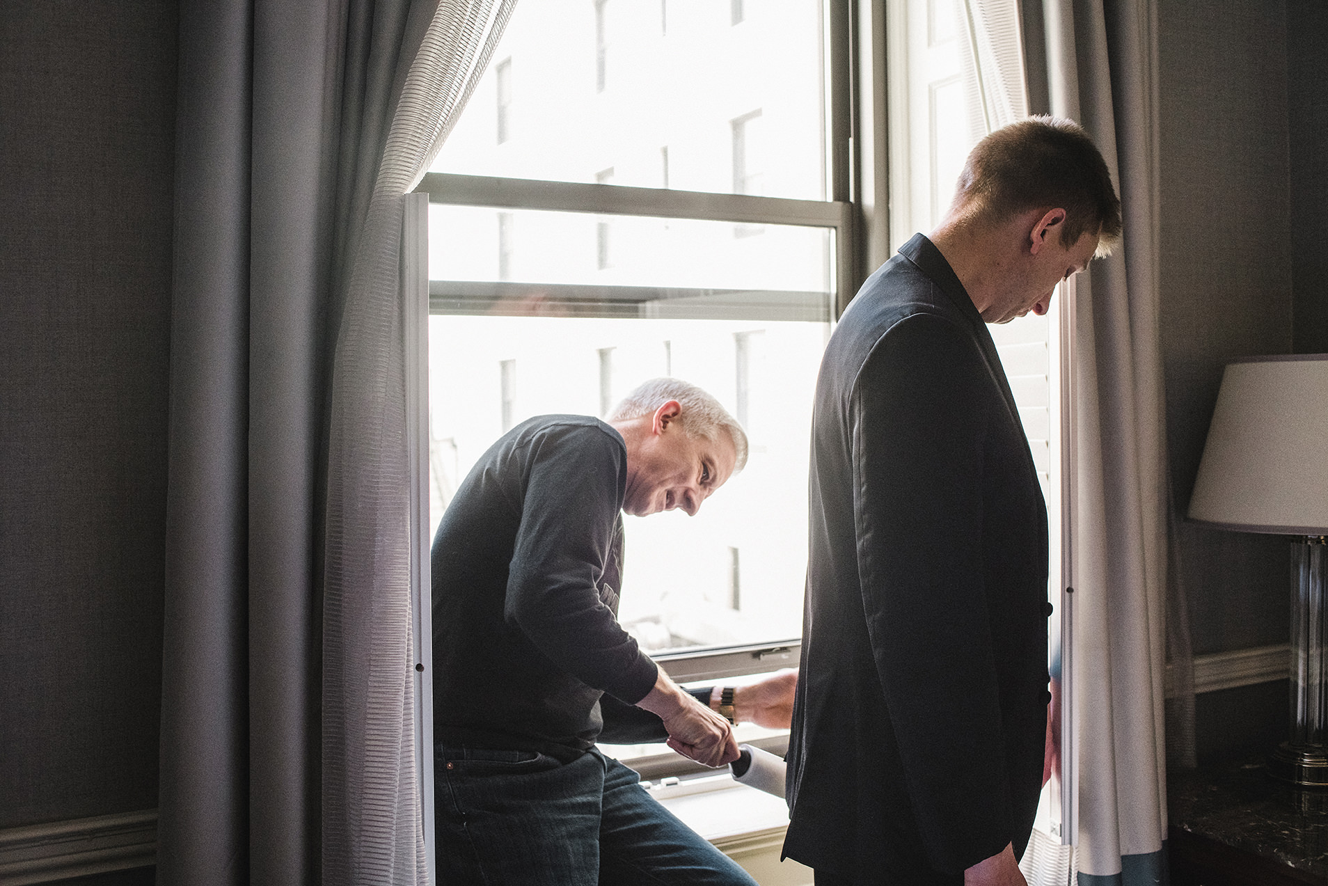 A documentary photograph of a father cleaning the lint of his sons suit before his Fairmont Copley Wedding