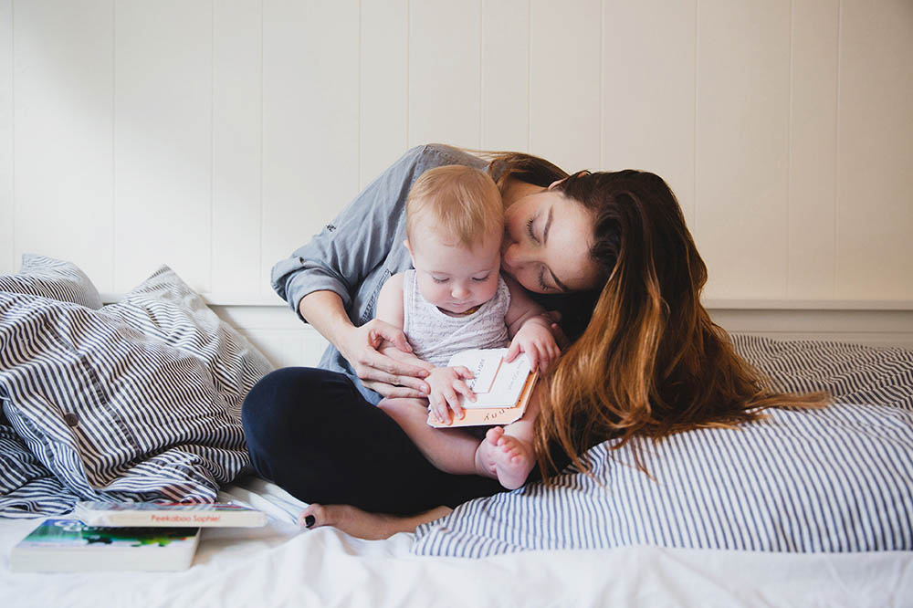 A documentary photograph of a mom kissing and cuddling her daughter as they read books during an in home family session in Boston