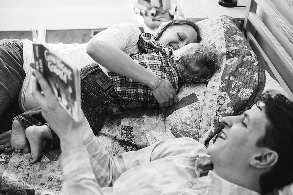 A documentary photograph of a mom snuggling her son as they read books in bed during an in home family session in Boston
