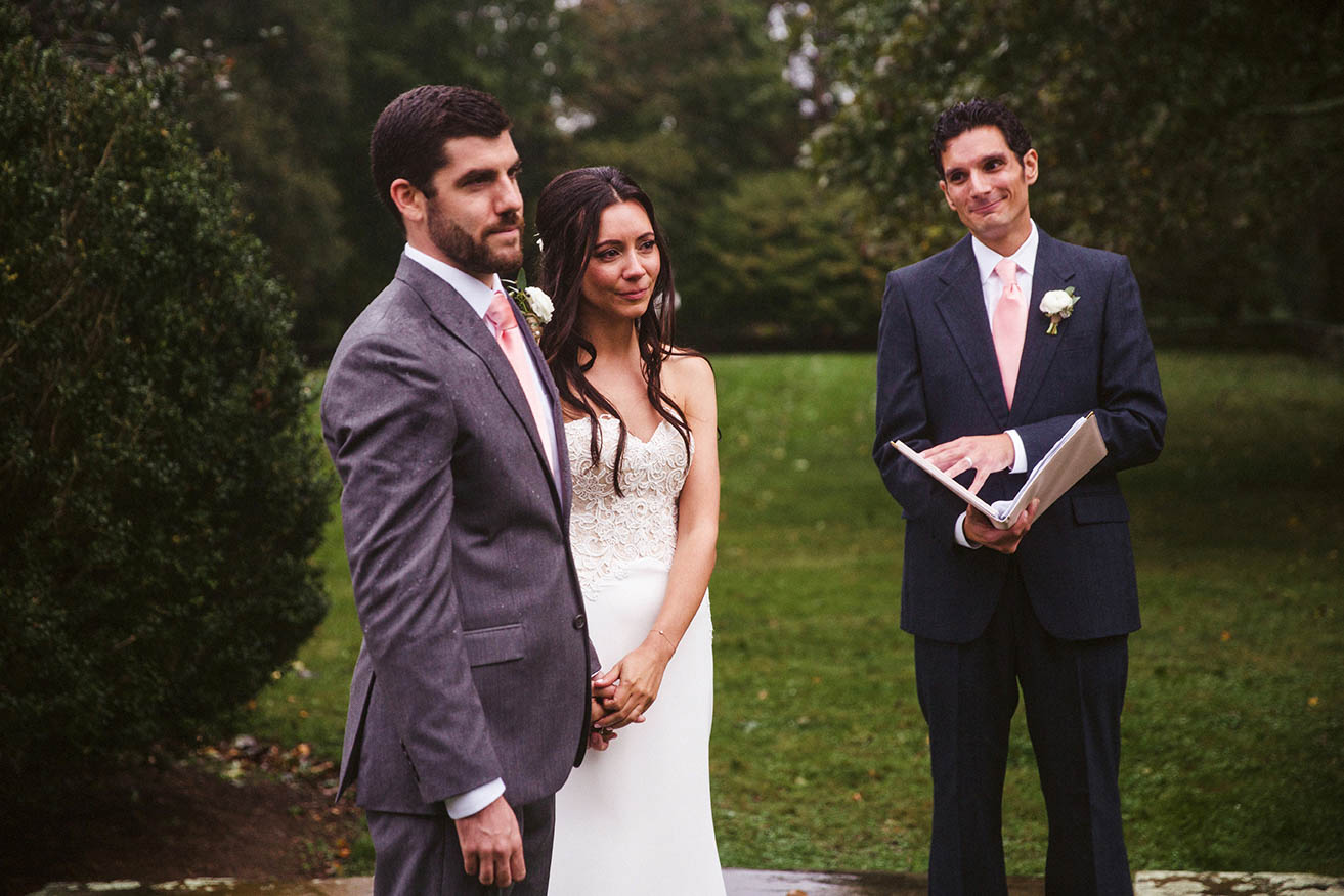 A documentary photograph of a bride and groom listening to a reading during their outdoor wedding ceremony in the rain