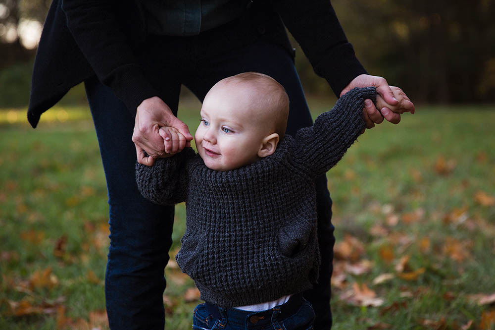 A documentary photograph of a baby walking with his mom during their family session at the Arnold Arboretum in Boston