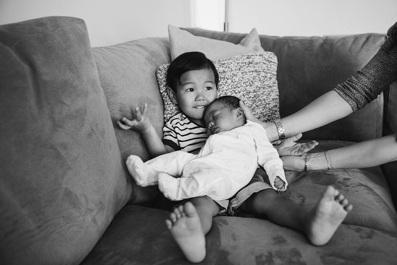 A documentary photograph of a boy trying to hold his newborn brother during an in home newborn family session in Boston's Jamaica Plain