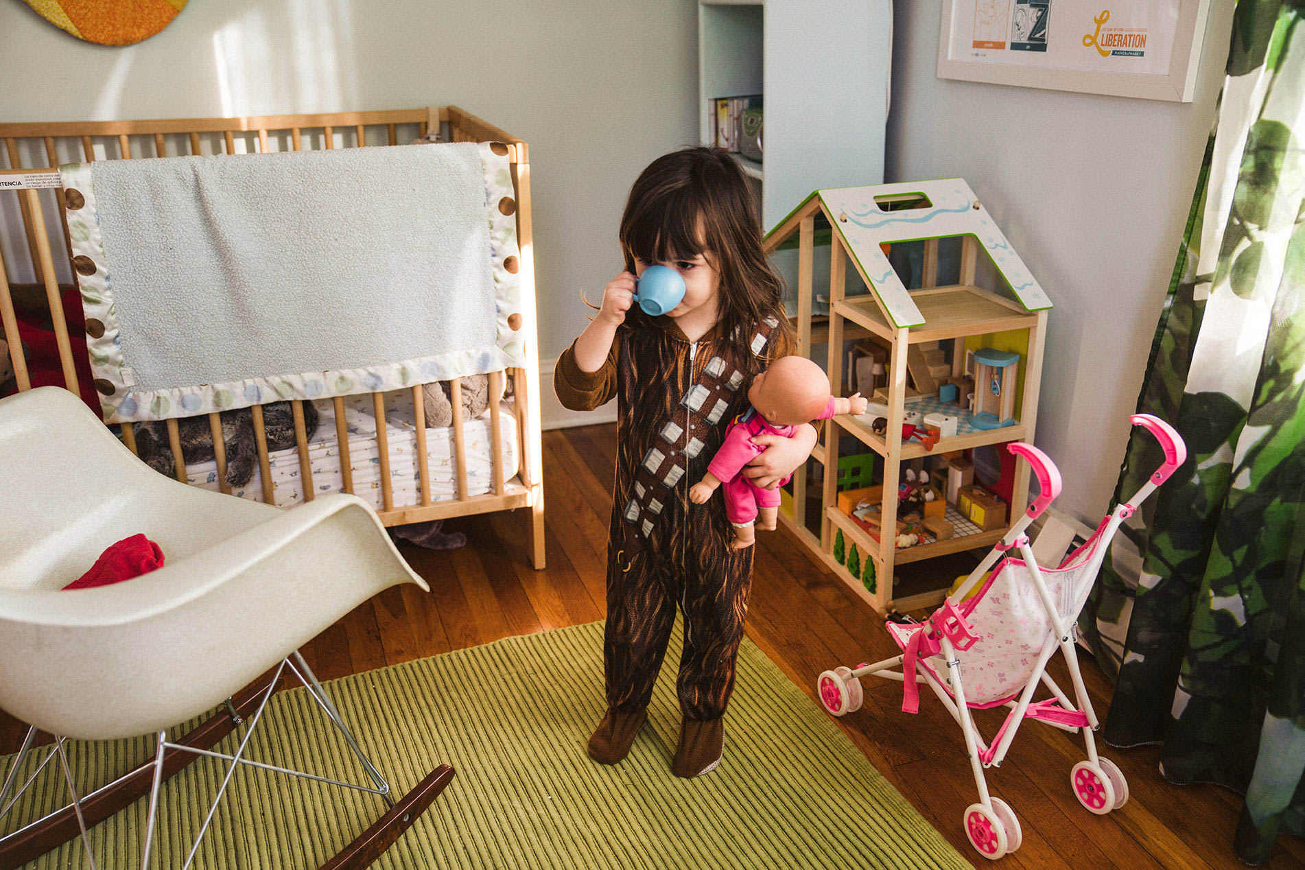 A documentary photograph of a toddler pretending to drink coffee while she holds a baby doll during an in home family session in Boston's Jamaica Plain