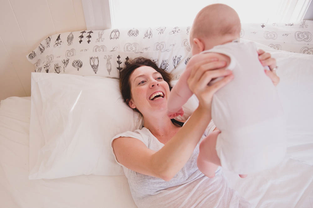 A documentary photograph of a mom playing with her baby in bed during an in home family session in Boston
