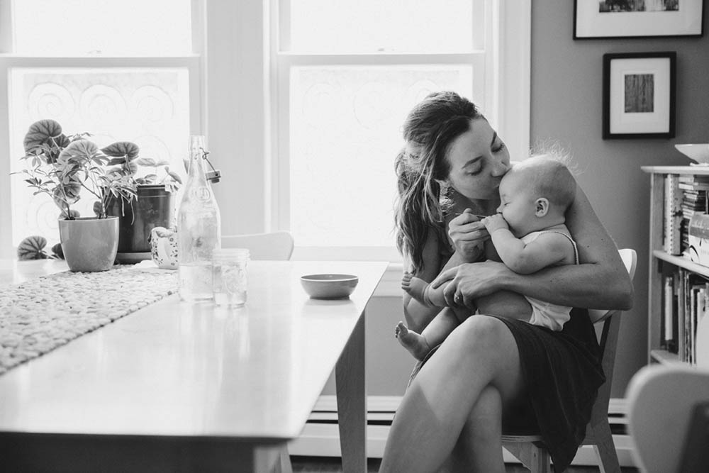 A documentary photograph of a mom kissing her baby while they eat breakfast during an in home family session in Boston