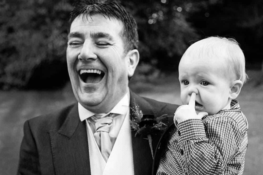 A documentary photograph of a grandfather laughing while holding her grandson who's picking his nose during a Boston wedding