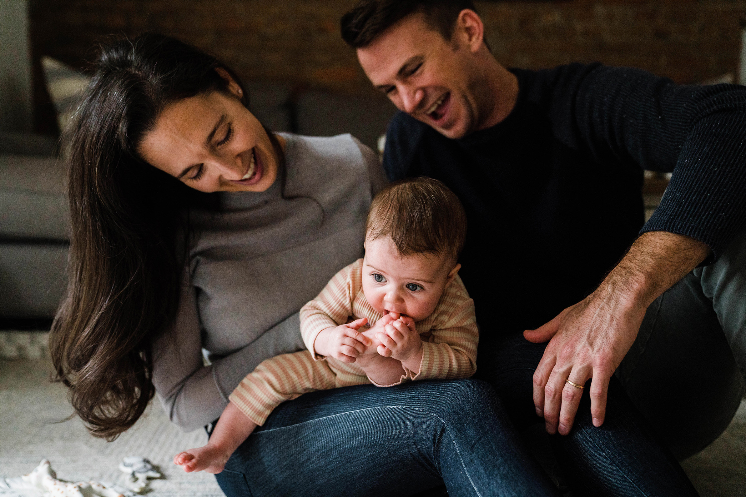 A documentary photograph of a parents laughing while their baby discovers her toes during an in home family session