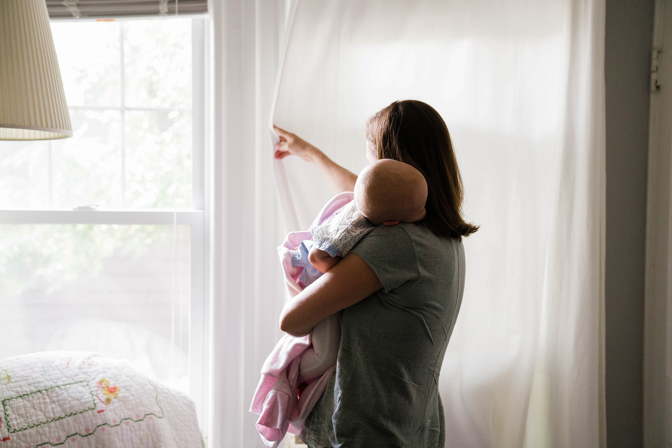 A documentary photograph of a mom closing the curtains while her baby sleeps on her shoulder during an in home family session in Boston
