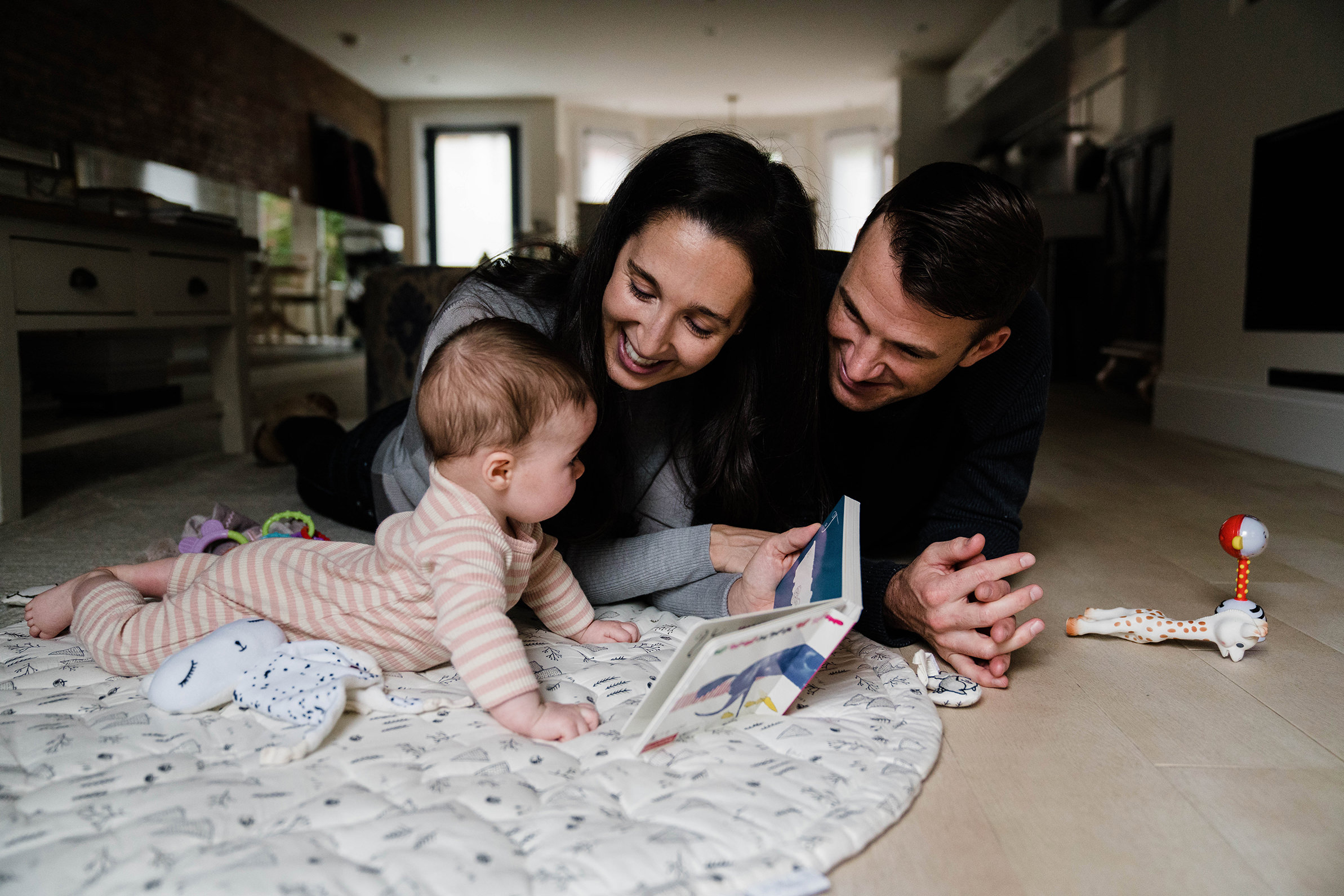 A documentary photograph of parents smiling at their baby while they read a book and have tummy time during their in home family session in Boston